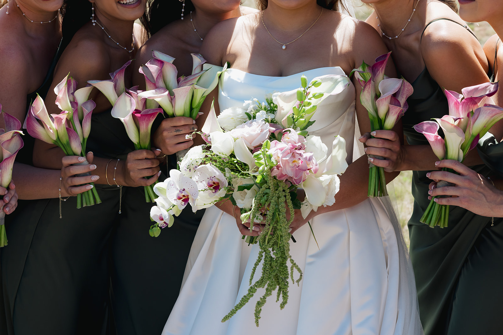 Bride in a strapless gown holding a cascading white and pink orchid bridal bouquet, surrounded by bridesmaids in dark green dresses holding calla lily bouquets.